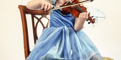 Mała dziewczynka o rudych włosach, ubrana w niebieską sukienkę grająca na skrzypcach.