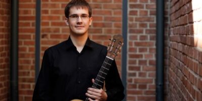 Młody mężczyzna w okularach ubrany w czarną koszulę. W lewej ręce trzyma gitarę klasyczną.