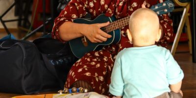 Małe dziecko siedzi tyłęm i słucha jak instruktorka, ubrana w czerwoną sukienkę, gra na małej gitarce