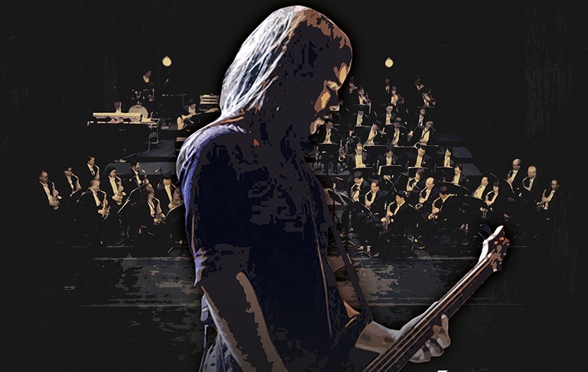Zdjęcie przedstawia długowłosego gitarzystę, a tle za nim muzycy orkiestry.