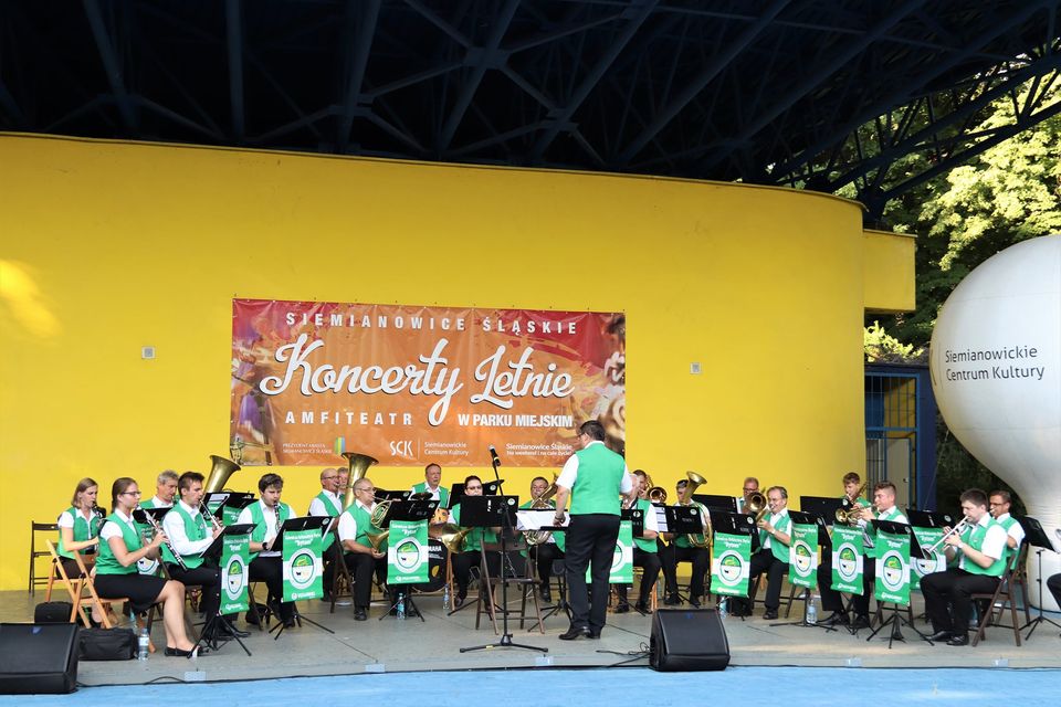 Na zdjęciu pełny skład Górniczej Orkiestry Dętej Bytom. Ubrani w biało zielone stroje muzycy grają na swoich instrumentach, na tle żółtej ściany amfiteatru. Foto Monika Bilska