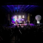Na zdjęciu widok ogólny amfiteatru podczas koncertu Rock Noc. Widać scenę z kolorowymi światłami, na niej zespół oraz ciemne sylwetki fanów i biały balon reklamowy SCK. Foto Monika Bilska
