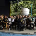 Widok boczny na siedzącą w strojach górniczych na scenie amfiteatru orkiestrę. W tle biały balon reklamowy SCK. Foto Monika Bilska