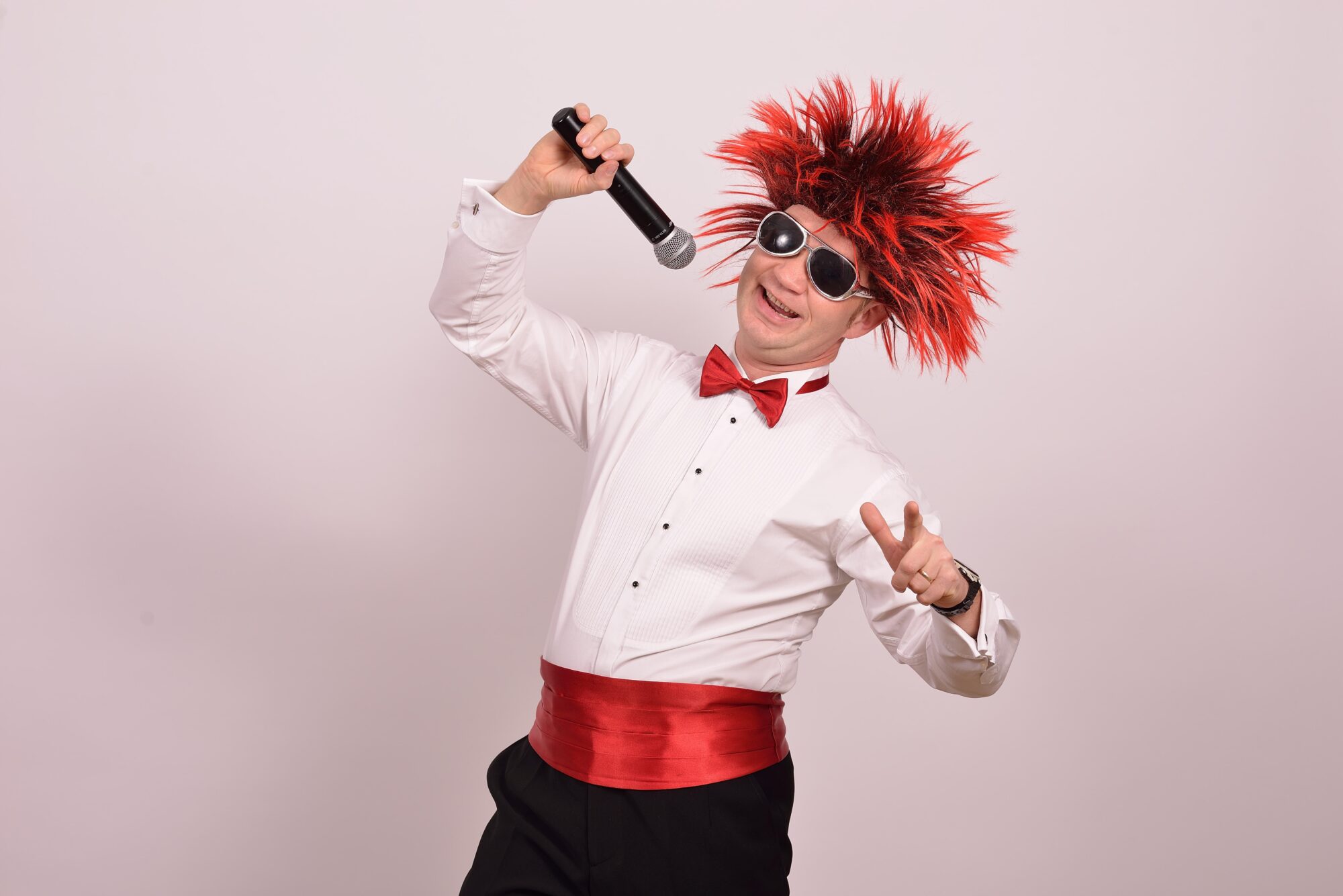 Wujek Kris czyli Krzysztof Kończyk ubrany w białą koszulę, czerwony pas i muszkę oraz w czerwonej peruce na głowie śpiewa do mikrofonu