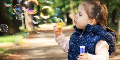 Zdjecie przedstawia dziewczynkę w niebieskim kropkowanym bezrękawniku i białej bluzeczce dmuchającą bańki mydlane na tle parkowej alejki.