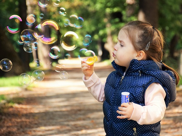 Zdjecie przedstawia dziewczynkę w niebieskim kropkowanym bezrękawniku i białej bluzeczce dmuchającą bańki mydlane na tle parkowej alejki.