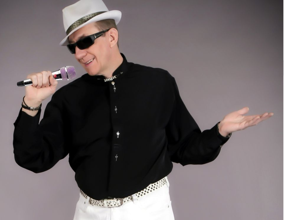 Artysta (Adi) ubrany w biały kapelusz z czarną lamówką, czarną koszulę, białe spodnie, śpiewający do mikrofonu znajdującego się w prawej dłoni artysty. Na nosie czarne okulary.