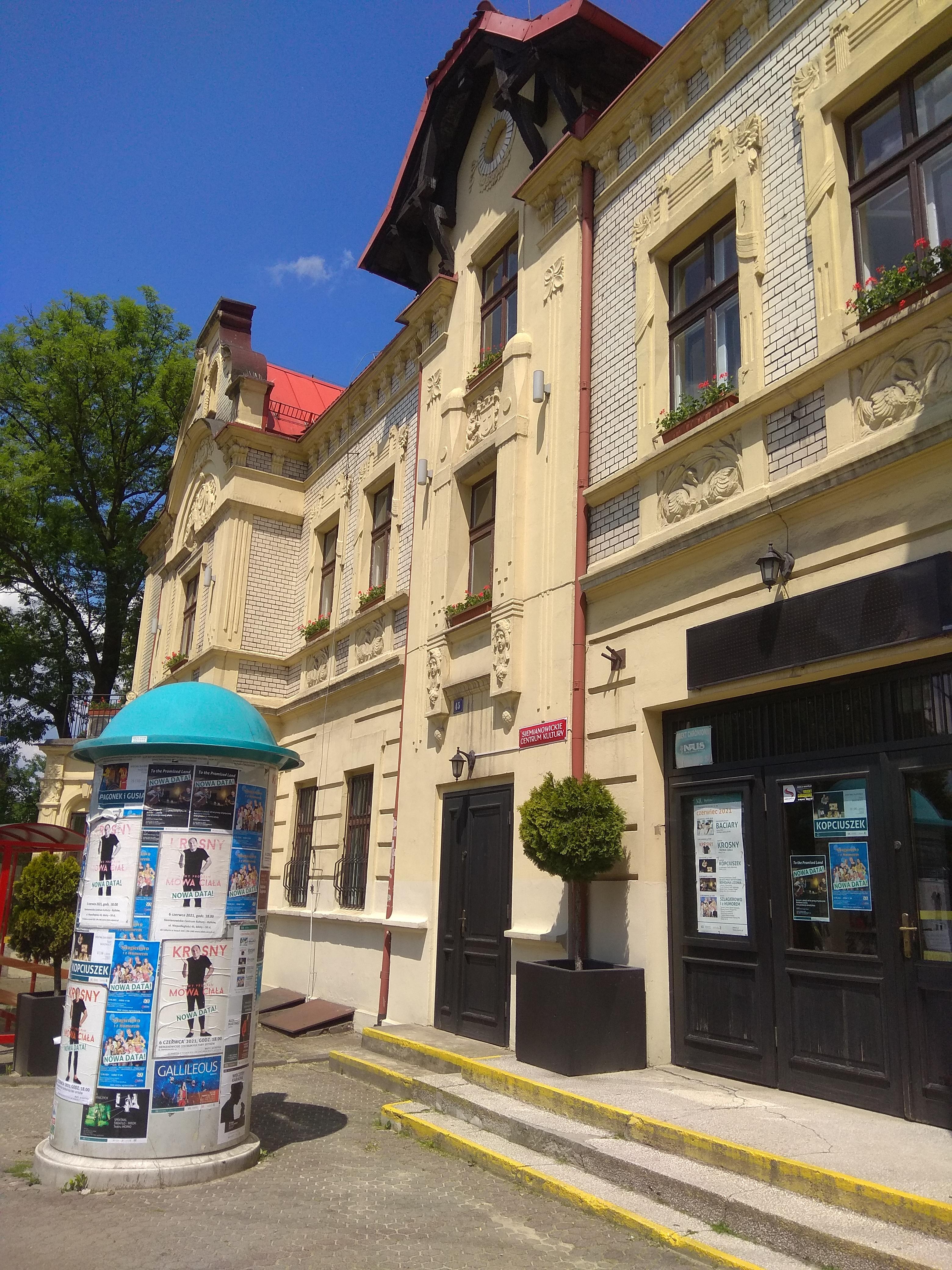 Kremowa fasada budynku SCK-Bytków, a przed nią słup ogłoszeniowy z plakatami reklamującymi wydarzenia