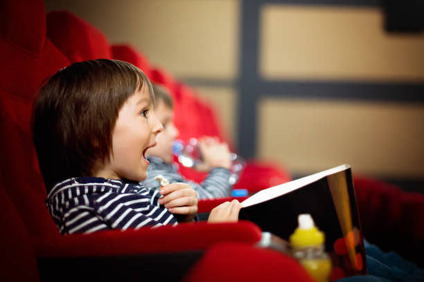 Uśmiechnięte dziecko siedzące na czerwonym fotelu w sali kinowej. W rękach trzyma kubełek z popcornem.