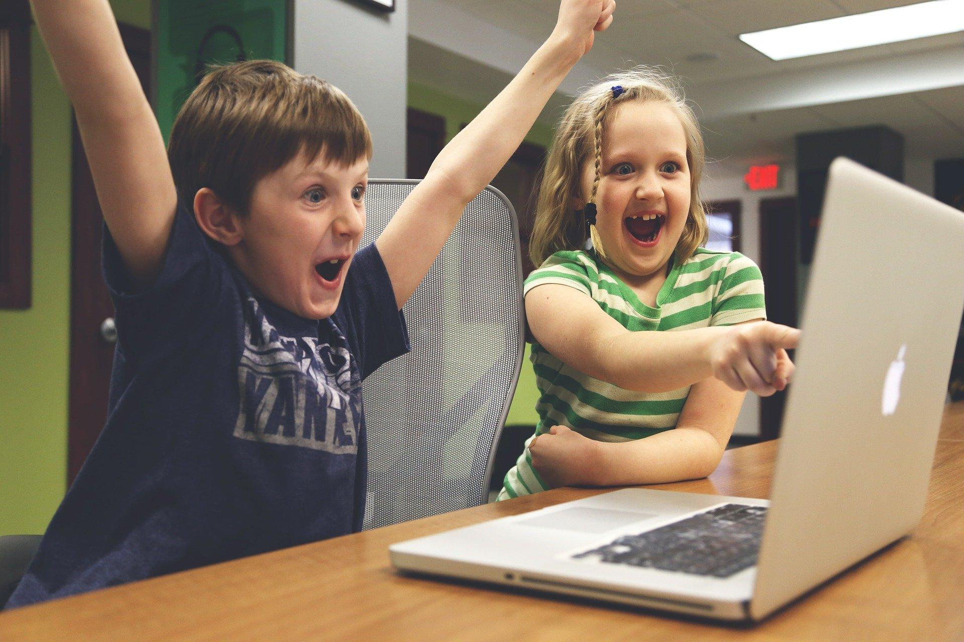 Dwoje dzieci: chłopeic i dzewczynka siedzą przy laptopie z rękoma w górze