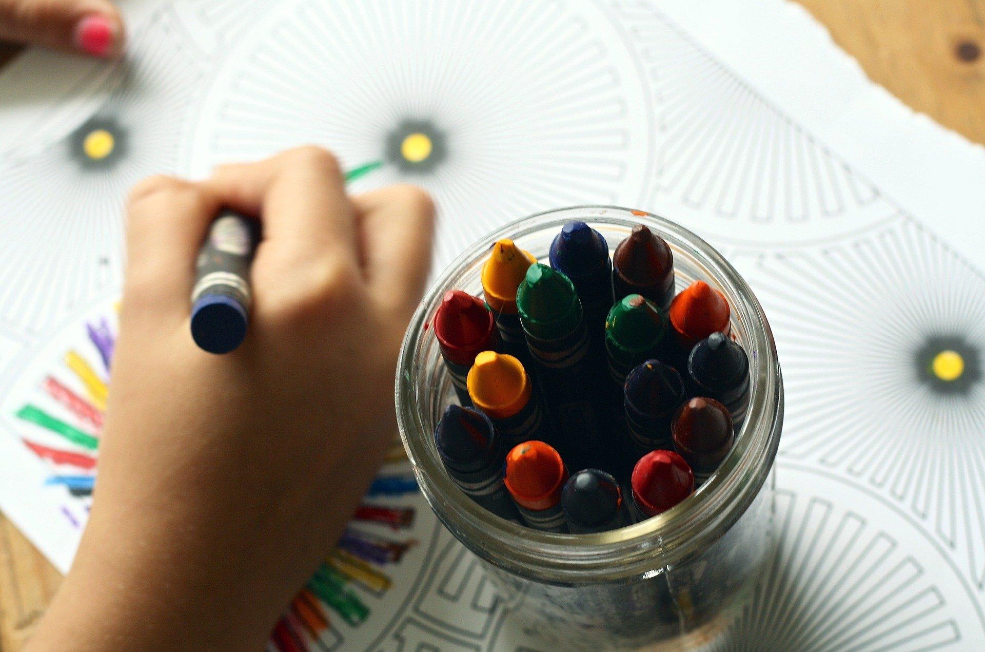 Na zdjęciu kredki świecowe w słoiczku oraz ręka dziecka rysująca nimi