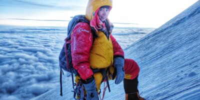 kobieta w grubym kombinezonie zimowym, na plecach plecak, stoi na zboczu ośniezonej góry, powyżej poziomu chmur