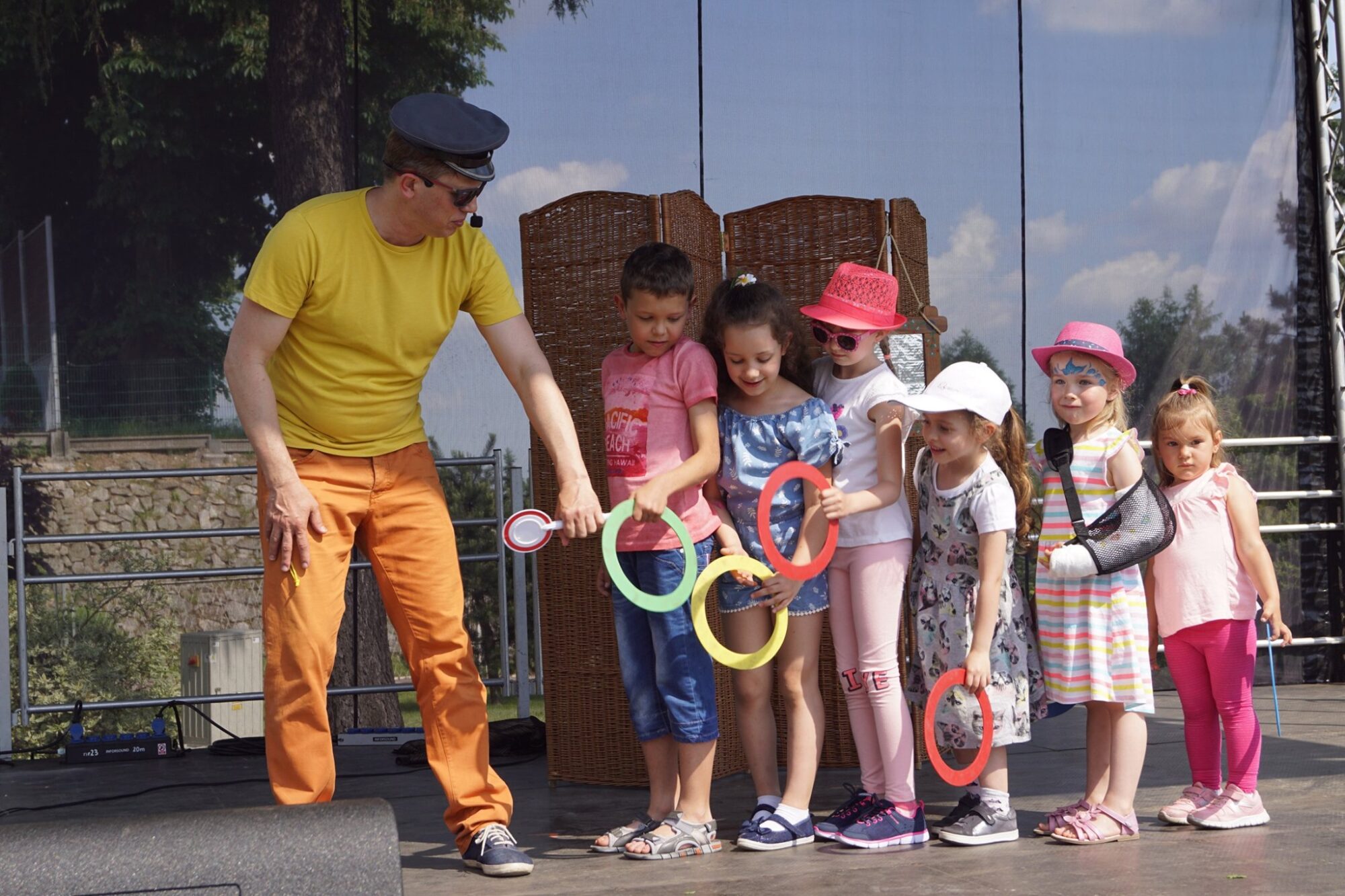 Na zdjęciu ubrany w kolorowy strój aktor bawi się z dziećmi, trzymającymi w rękach kolorowe kółka