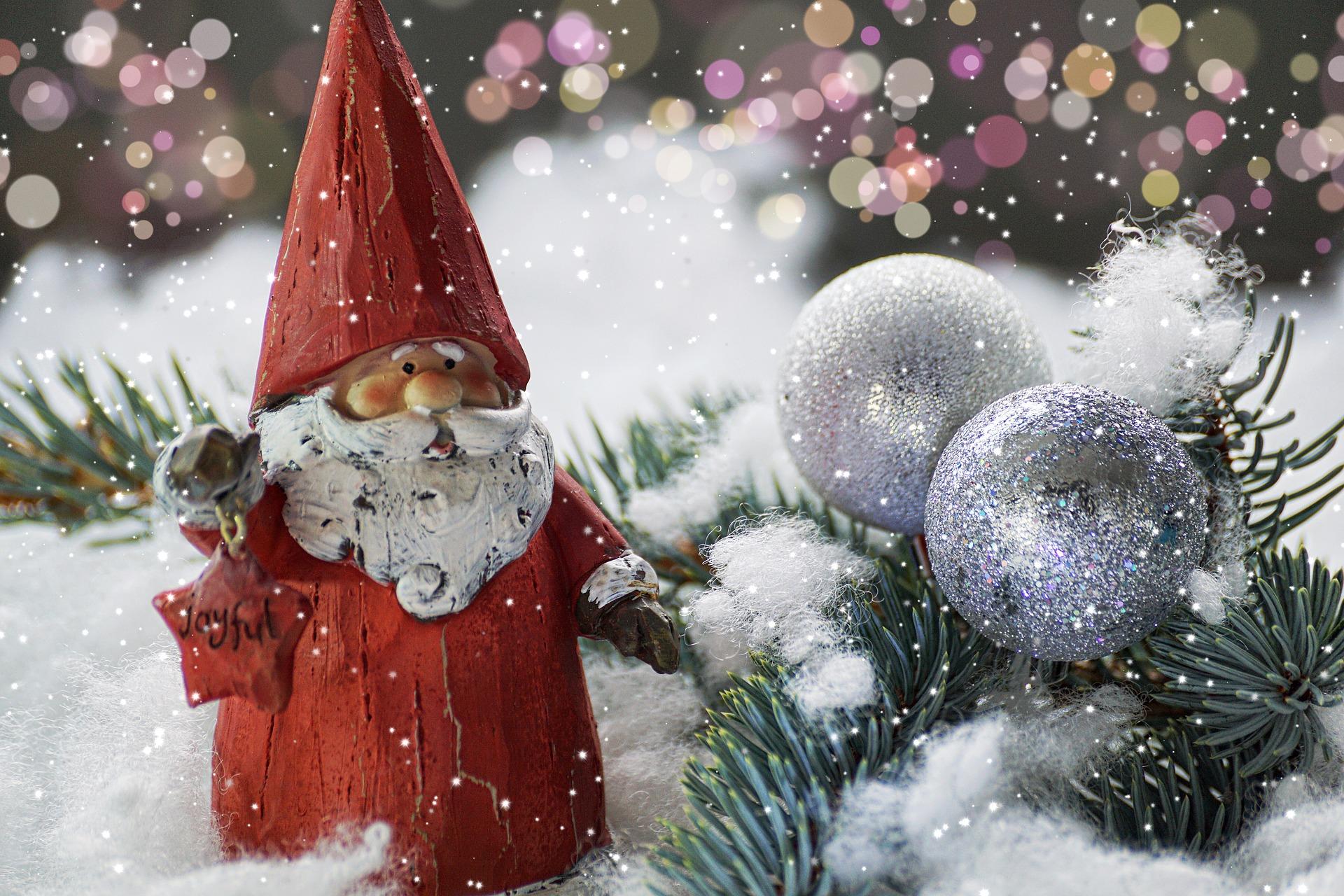 figurka mikołaja z białą, długą brodą, w czerwonym ubraniu i czapce stoi w sniegu obok gałązek przybranych bombkami