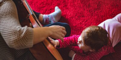 Na czerwonym dywanie leży male dziecko i wyciąga rękę w stronę gitary, na któej gra młoda kobieta